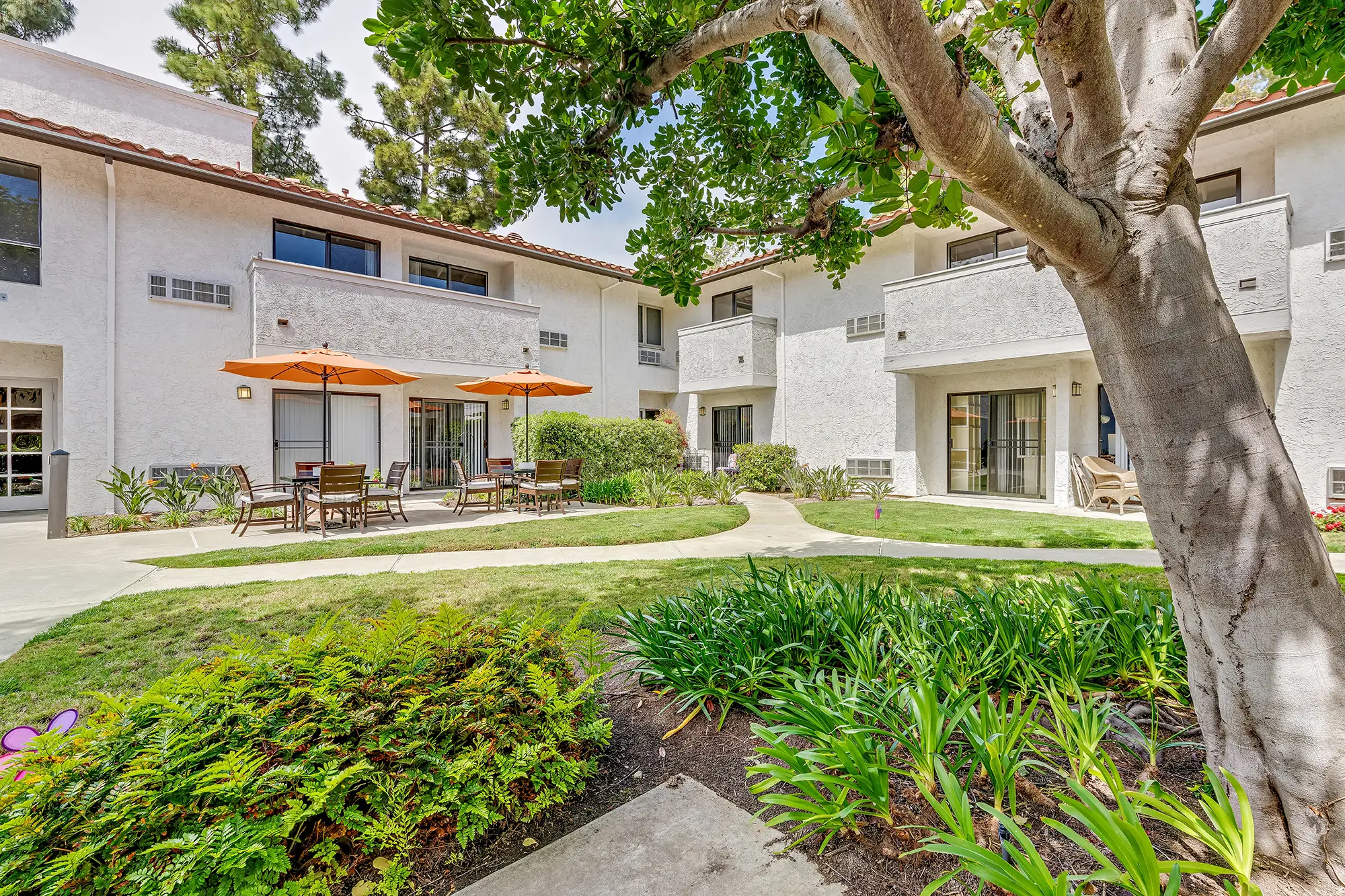 An exterior view of 3 tables with 11 chairs and two umbrellas outside of  a 55+ apartment senior living community